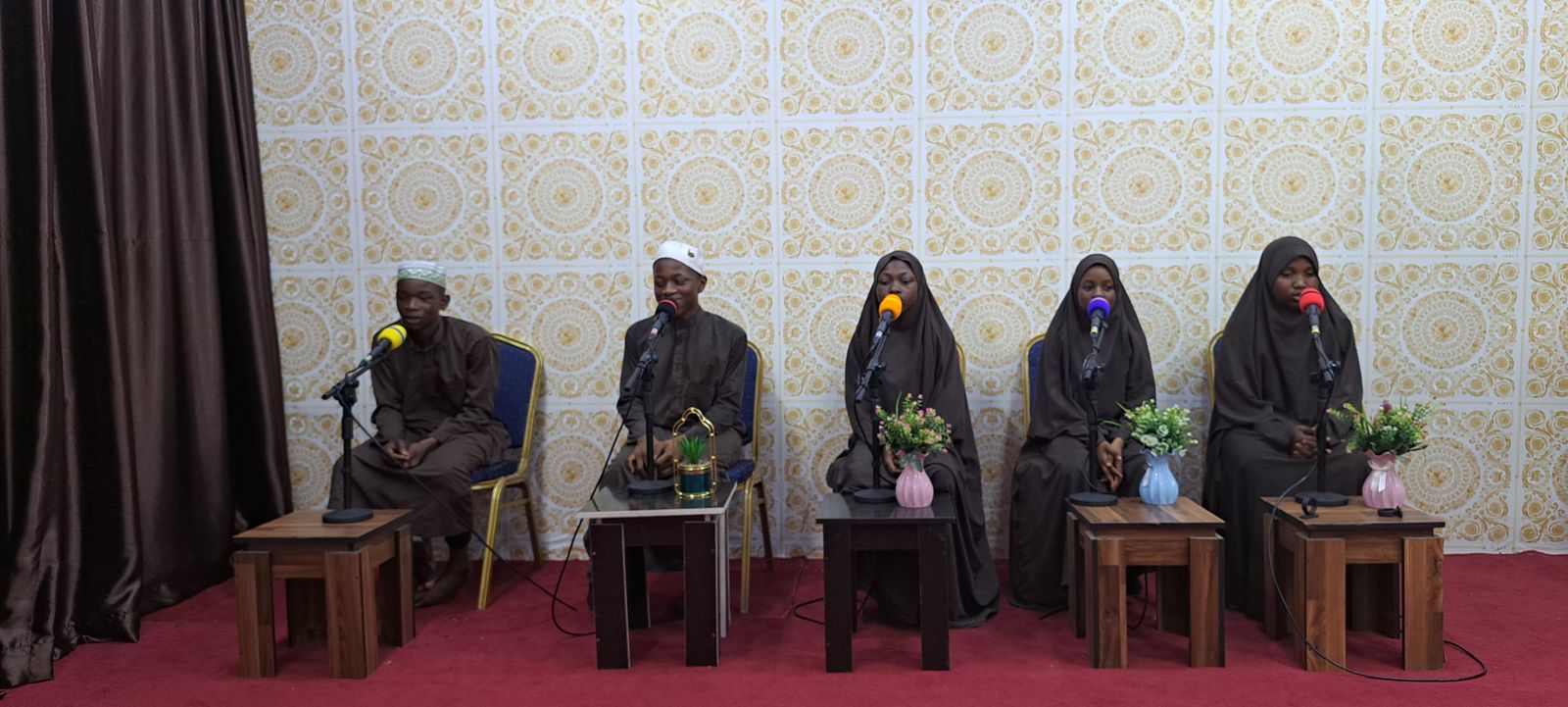 Qur'an College students seated on stage with microphones during a Qur'an recitation presentation